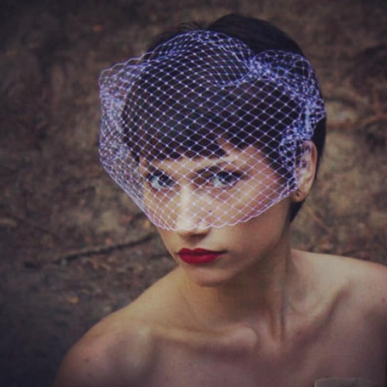 May include: A woman with short, dark hair wears a white birdcage veil with a fine mesh pattern. She is wearing red lipstick and has a neutral expression.