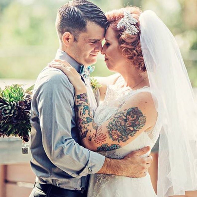 Puede incluir: Una pareja en atuendo de boda, la novia lleva un vestido blanco y velo, el novio lleva una camisa azul claro y una corbata de lazo. Ambos tienen tatuajes coloridos en sus brazos.