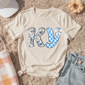Puede incluir: Camiseta color crema con las letras "KY". La "K" está decorada con un estampado floral en tonos azules y grises, mientras que la "Y" tiene un diseño a cuadros azul y blanco. Un lazo blanco está en la "Y".