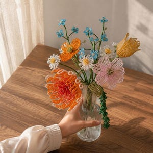 May include: A bouquet of handcrafted beaded flowers in a clear glass vase. The arrangement features orange, pink, yellow, white, and light blue blossoms with green beaded stems and leaves, showcasing intricate wire and bead construction throughout.