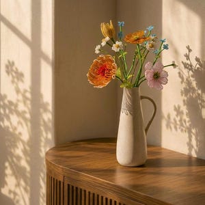 May include: A tall, off-white ceramic pitcher vase with a single handle, containing a bouquet of handcrafted beaded flowers in shades of orange, yellow, pink, white, and blue, with green stems and leaves arranged in a decorative display.
