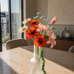 May include: A bouquet of handcrafted beaded flowers in a clear, vertically ribbed glass vase. The arrangement features vibrant red poppies with dark centres, soft pink lilies, white daisies with yellow centres, and small blue blossoms on green stems.