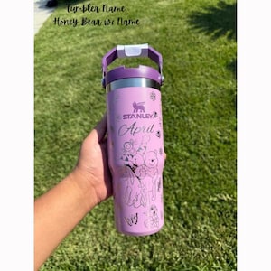 Peut inclure: Un gobelet Stanley lavande avec une poignée et un couvercle violets. Le gobelet présente un motif Winnie l'ourson avec le mot "April" et le logo Stanley. Le texte "Tumbler Name" et "Honey Bear w/ Name" sont en haut.