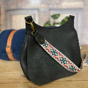May include: Black leather crossbody bag with a patterned strap featuring red, turquoise, and white geometric designs. The bag has a textured surface and a gold-colored clasp. A blue and tan bag is in the background.