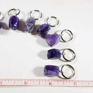 May include: Seven amethyst stone keychains with silver key rings. The stones are a deep purple color and have a natural, rough texture. The keychains are arranged in a row on a white surface with a ruler for scale.