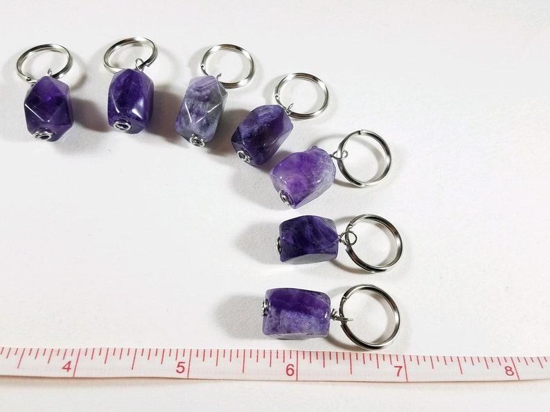 May include: Seven amethyst stone keychains with silver key rings. The stones are a deep purple color and have a natural, rough texture. The keychains are arranged in a row on a white surface with a ruler for scale.