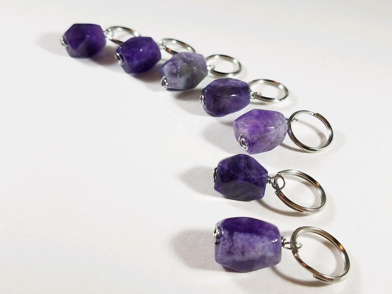 May include: Seven amethyst gemstone stitch markers with silver rings. The amethyst stones are various shades of purple and have a faceted, irregular shape.