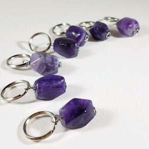 May include: Six amethyst crystal stitch markers with silver key rings. The amethyst crystals are various shades of purple and have a natural, rough texture.