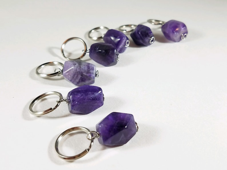 May include: Six amethyst crystal stitch markers with silver key rings. The amethyst crystals are various shades of purple and have a natural, rough texture.