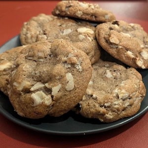 May include: A plate of freshly baked cookies. The cookies are a light brown color, with visible chunks of white chocolate and nuts. They are arranged on a dark gray plate, set against a red surface.