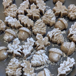 May include: A dark grey plate with leaf and acorn-shaped treats dusted with icing sugar. The treats are light brown and arranged in a random pattern, creating a rustic, autumnal theme.