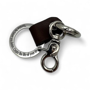 May include: A silver-toned keychain with a brown leather strap. The keychain has a large ring with the engraved text "Envision the future you desire. Create the life of your dreams." A lobster clasp and a smaller ring are also present.