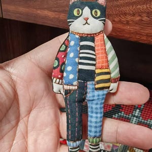 May include: A fabric cat doll featuring a patchwork design with various patterns including polka dots, stripes, and plaid in colors of black, white, blue, orange, and green. The cat has large yellow eyes, a small pink nose, and visible stitching.