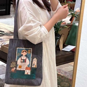 Op de afbeelding: Een donkergrijze tote bag met een kleurrijke illustratie van een meisje, huizen en een boom. De tas wordt over de schouder gedragen en heeft een rechthoekig ontwerp met een lange band.