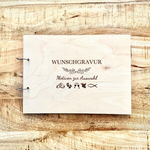 Könnte beinhalten: Ein helles Holzbuch mit Metallbinderingen. Der Einband zeigt eingravierten Text und florale Akzente sowie verschiedene Symbole. Der Text lautet "WUNSCHGRAVUR" und "Motiven zur Auswahl".
