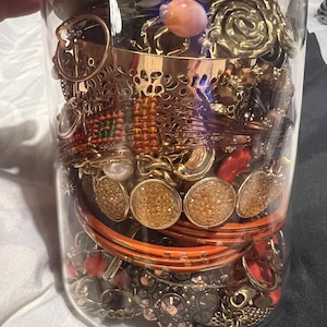 May include: A glass jar containing an assortment of jewellery, such as bracelets, rings, and necklaces. The jewellery is mainly gold and orange, with some pieces incorporating gemstones and pearls. The jar is topped with a wooden lid.
