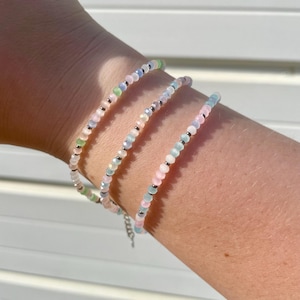 May include: Three delicate beaded bracelets in pastel shades of pink, blue, green, and white. The bracelets are strung with small, round beads and silver-coloured spacers. The bracelets are worn on a wrist.