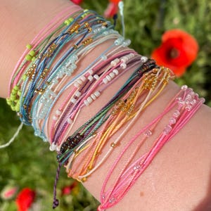 May include: A selection of colourful beaded bracelets in shades of pink, green, blue, and gold. The bracelets are made of string and small beads, and are stacked. The bracelets are a mix of solid colours and patterns.