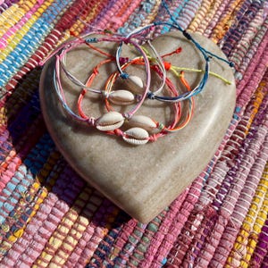 May include: A collection of colourful friendship bracelets, each adorned with cowrie shells, rests on a heart-shaped stone. The bracelets feature threads in pink, orange, blue, and yellow. The stone sits on a multi-coloured woven textile.