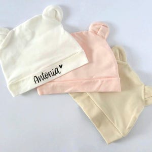 Könnte beinhalten: Drei Baby-Mützen in Creme, Rosa und Beige. Jede Mütze hat kleine, abgerundete Ohren oben. Die cremefarbene Mütze trägt den Namen "Antonia" in schwarzer Schrift. Die Mützen sind aus weichem Stoff und haben einen Umschlag.