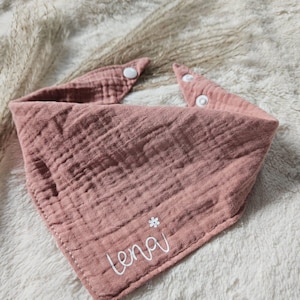 Könnte beinhalten: Ein rosa Mull-Baby-Bandana-Lätzchen mit weißen Druckknöpfen und dem Namen "Lena", der in Weiß mit einer kleinen weißen Blume gestickt ist.