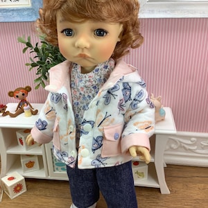 May include: A doll with light brown curly hair wearing a white jacket with butterfly and dragonfly designs, a floral print shirt, and blue jeans.