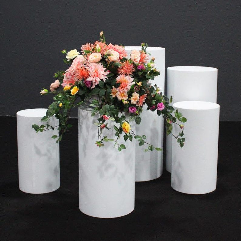 May include: A floral arrangement with pink, peach, yellow, and purple flowers and green foliage sits atop a white cylindrical pedestal. Several other white pedestals of varying heights are arranged around the main one, creating a display.