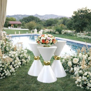 May include: Three white hourglass-shaped pedestal tables with gold-toned circular accents at the center, arranged in a cluster. A large, rounded floral arrangement featuring orange, pink, and white roses sits atop the central table.
