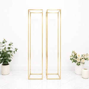 May include: Two tall, slender, rectangular display stands constructed from thin, metallic gold-finished metal rods. Each stand features an open-frame design with a square base and a matching square top, creating a hollow, vertical structure.