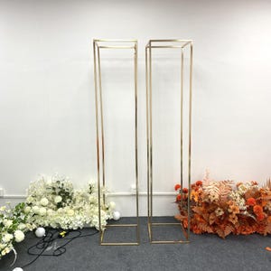 May include: Two tall, gold metal rectangular stands. The stands are open at the top and bottom, with a square base. The stands are placed in front of white and orange floral arrangements.