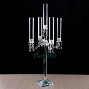 May include: A clear crystal candelabra with five candle holders and a square base. The candelabra is decorated with crystal drops.