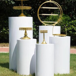 May include: An assortment of gold-colored serving stands and display shelves on white cylindrical pedestals. The stands vary in design, including tiered trays and a circular shelf. The setting is outdoors on a grassy area with green foliage in the background.