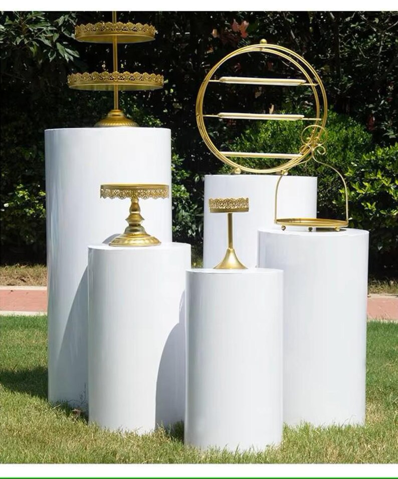 May include: An assortment of gold-colored serving stands and display shelves on white cylindrical pedestals. The stands vary in design, including tiered trays and a circular shelf. The setting is outdoors on a grassy area with green foliage in the background.