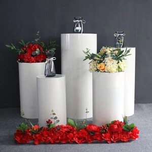 May include: A display of white cylindrical pedestals of varying heights, adorned with floral arrangements and small glass vases. Red and yellow flowers are featured, creating a visually appealing contrast against the white surfaces. The arrangement is set against a dark gray backdrop.