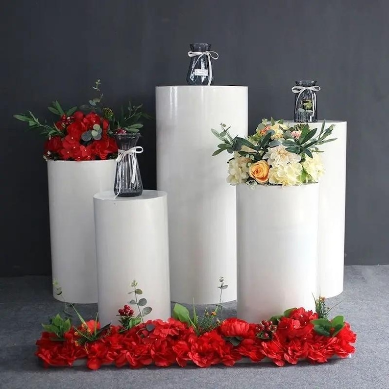 May include: A display of white cylindrical pedestals of varying heights, adorned with floral arrangements and small glass vases. Red and yellow flowers are featured, creating a visually appealing contrast against the white surfaces. The arrangement is set against a dark gray backdrop.