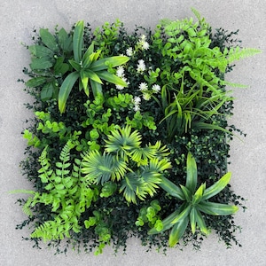 May include: A square artificial plant panel featuring a variety of green foliage. The panel includes different types of leaves, including ferns, broad leaves, and small, round leaves. Small white flowers are interspersed throughout the panel, adding a touch of contrast.