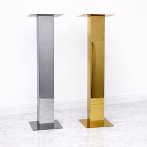 May include: Two tall, slender, rectangular display pedestals with a reflective metallic finish. One pedestal is silver-toned, and the other is gold-toned. Each features a flat square base and a matching flat square top connected by a vertical column.