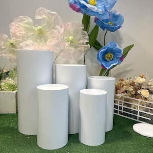 May include: A collection of white cylindrical pedestals of varying heights, arranged on a green surface. The pedestals are smooth and evenly colored. Large, artificial flowers in shades of blue and pink are in the background, suggesting a display or event setting.