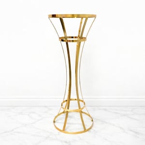 May include: A tall, gold metal decorative stand with a round top and a flared, curved design. The stand features a circular base and a mid-section ring, creating an elegant, open structure. Ideal for displaying plants or decorative items.