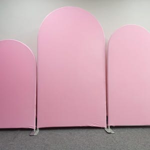 May include: Three pink arched backdrop stands of varying sizes. The stands are made of a metal frame and covered with a smooth, solid pink fabric. The backdrop stands are designed for events and photo shoots.