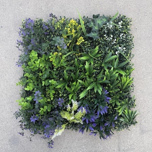 May include: A square artificial plant panel featuring a variety of green, purple, and yellow faux foliage. The panel includes different textures and shades of green, with accents of purple flowers and yellow buds. The overall effect is a vibrant, natural-looking display.