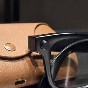 May include: Black smart glasses featuring a prominent circular button on the temple frame, positioned next to a tan textured carrying case with a metallic snap closure and a visible charging port at the bottom edge of the case.