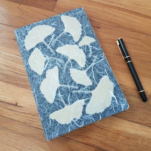 May include: A dark blue notebook with a textured cover featuring white paper shapes. The notebook is lying on a wooden surface with a black pen next to it.