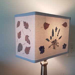 May include: A rectangular lampshade with a light beige fabric, adorned with pressed leaves and blue floral accents. The top and bottom edges are trimmed with a light blue border. The lamp base is a brushed metal finish.