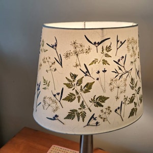May include: A lampshade with a botanical design. The beige lampshade features green leaves and purple flowers. The cylindrical lampshade tapers towards the top, with a silver metal base. The design includes pressed flowers and foliage.