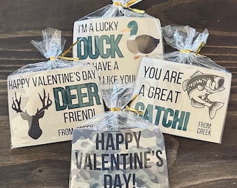 May include: Four Valentine's Day cards with a hunting and fishing theme. The cards are packaged in clear cellophane bags with gold ribbon. The cards feature a deer, a duck, a fish, and camouflage print. The text on the cards reads "I'm a lucky duck, have a deer like you", "You are a great catch", and "Happy Valentine's Day!"