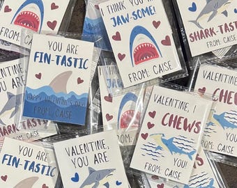 May include: A collection of colorful Valentine's Day cards featuring shark illustrations and puns. The cards have messages like "You are Fin-Tastic", "I think you are Jaw-some!" and "Valentine, I Chews You!"