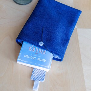 May include: A blue book sleeve with a button closure, holding a book titled "LES FILLES" by Anne Jacobs. The sleeve is made of textured fabric and has a striped bookmark.