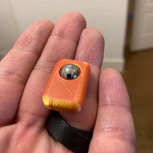 Puede incluir: Juguete fidget naranja y amarillo con un rodamiento de bolas plateado en el centro. El juguete tiene una superficie texturizada y se sostiene sobre un anillo negro.