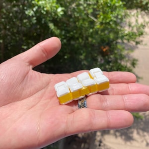 Può includere: Un piccolo giocattolo bianco e giallo che assomiglia a una fila di auto in miniatura. Il giocattolo è tenuto in una mano, con uno sfondo verde sfocato di fogliame. Le auto giocattolo hanno la parte superiore bianca e la base gialla.